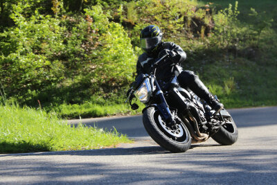 Ein schwarzes Motorrad mit einem Mann darauf biegt in eine Kurve. Im Hintergrund sieht man frisch gewachsenes Frühlingsgras.
