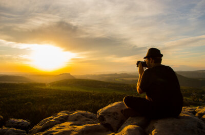 Johnny sitzt mit Kamera und fotografiert einen Sonnenuntergang auf einem Berg.