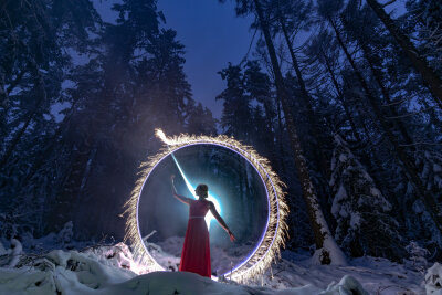 Eine Frau im rotem langen Kleid steht im Schnee. Hinter ihr der Winterwald und ein weißer Lichtschein.