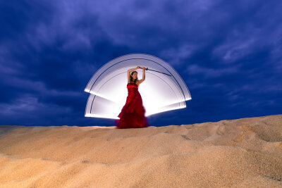 Eine Frau mit rotem langen Kleid steht auf einer Sanddüne. Hinter ihr ein weißer Lichtschein und der Nachthimmel.