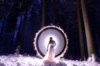 Eine Frau im weißen Kleid steht im Schnee. Hinter ihr der Winterwald und ein weißer Lichtschein.