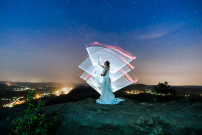 Frau im weißen Kleid und weißer Lichtkunst hinter ihr steht auf einem Berg. Dahinter die Lichter der Stadt.