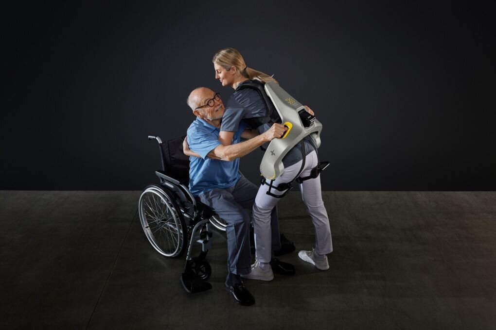 Intelligente Kraftanzüge sollen Pflegekräfte unterstützen - Eine Frau hebt mit Hilfe eines intelligenten Kraftanzugs der Firma German Bionic einen Mann aus einem Rollstuhl.
