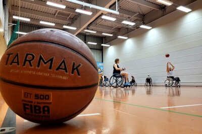 Ein Basketball liegt in der Halle. Im Hintergrund sieht man viele Menschen in Rollstühlen Basketball spielen.