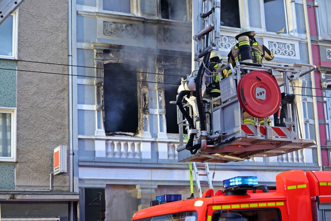 Solingen: Familie mit zwei Kindern stirbt bei Brand - Feuerwehrmänner stehen vor dem Mehrfamilienhaus in Solingen; Rauch dringt aus einem der Fenster.