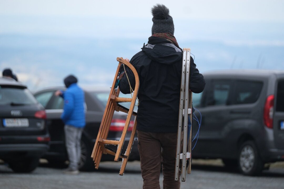 Tourismusministerium: Erfolgreicher Winter für Sachsen - Ein Mann trägt auf dem Fichtelberg im Erzgebirge zwei Rodelschlitten über einen Parkplatz.